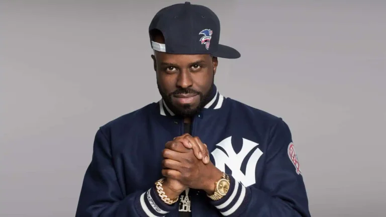 DJ Funk Flex wearing a navy blue New York Yankees jacket and cap, clasping his hands with gold jewelry visible.
