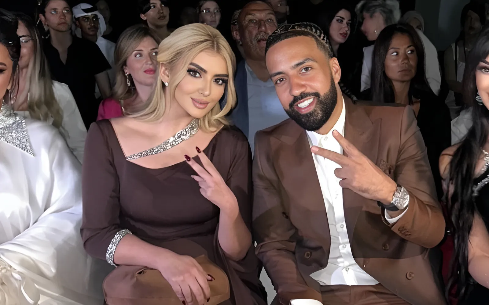 French Montana sitting with Sheikha Mahra at a fashion show, both smiling and posing for the camera.