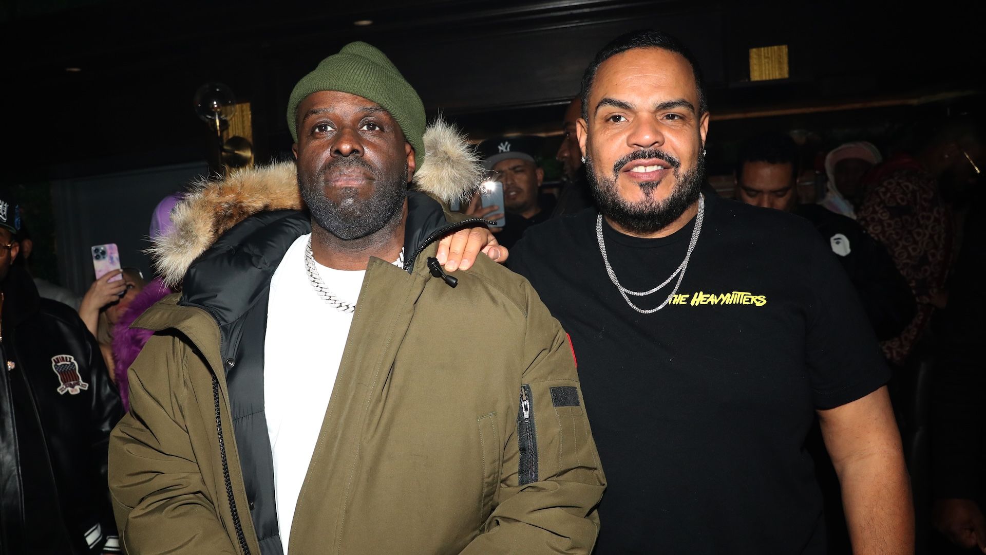 Funk Flex wearing a green beanie and parka jacket standing next to DJ Enuff in a black “The Heavy Hitters” t-shirt at a hip-hop event.