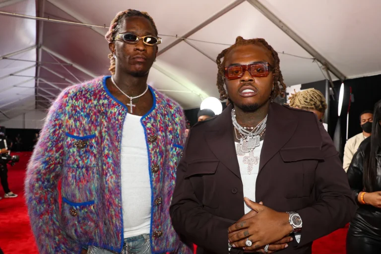 Young Thug wearing a colorful cardigan and sunglasses standing next to Gunna in a dark suit with layered diamond chains on the red carpet.