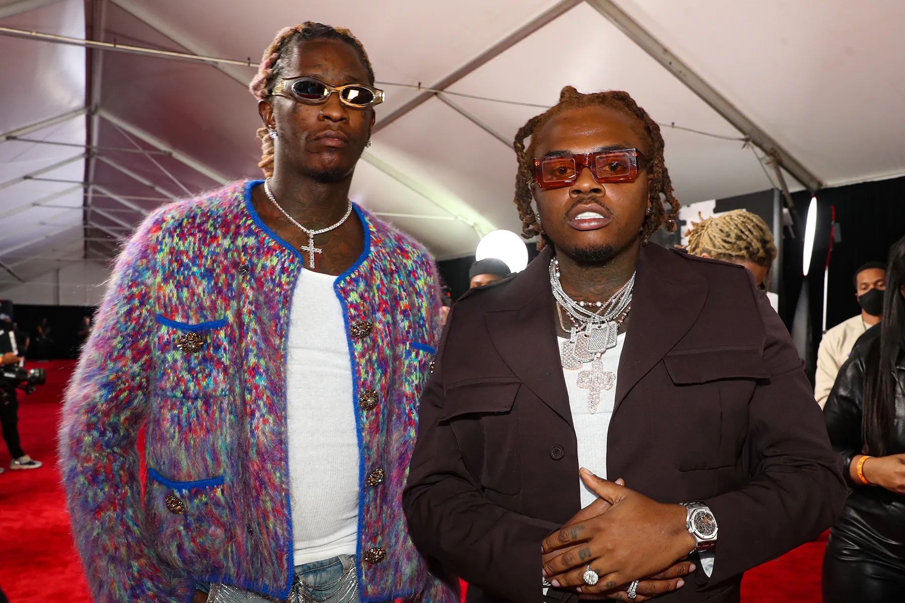 Young Thug wearing a colorful cardigan and sunglasses standing next to Gunna in a dark suit with layered diamond chains on the red carpet.