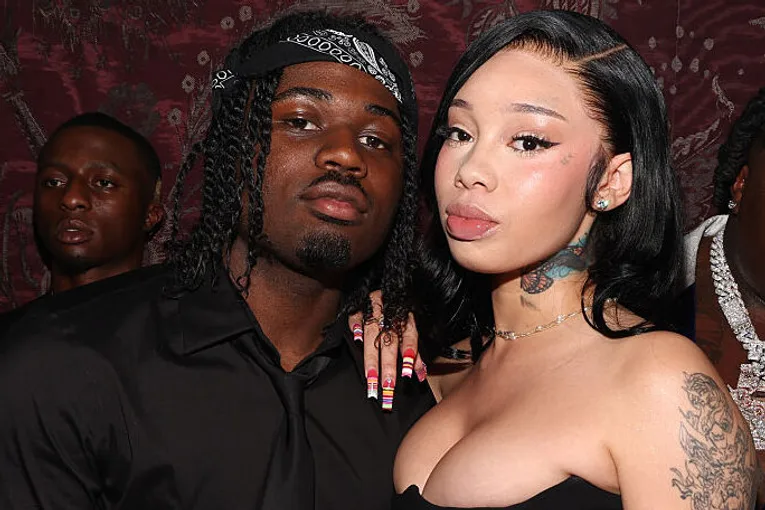 Rapper 4Batz wearing a black shirt and bandana posing beside his girlfriend Anycia in a strapless black dress with tattoos.