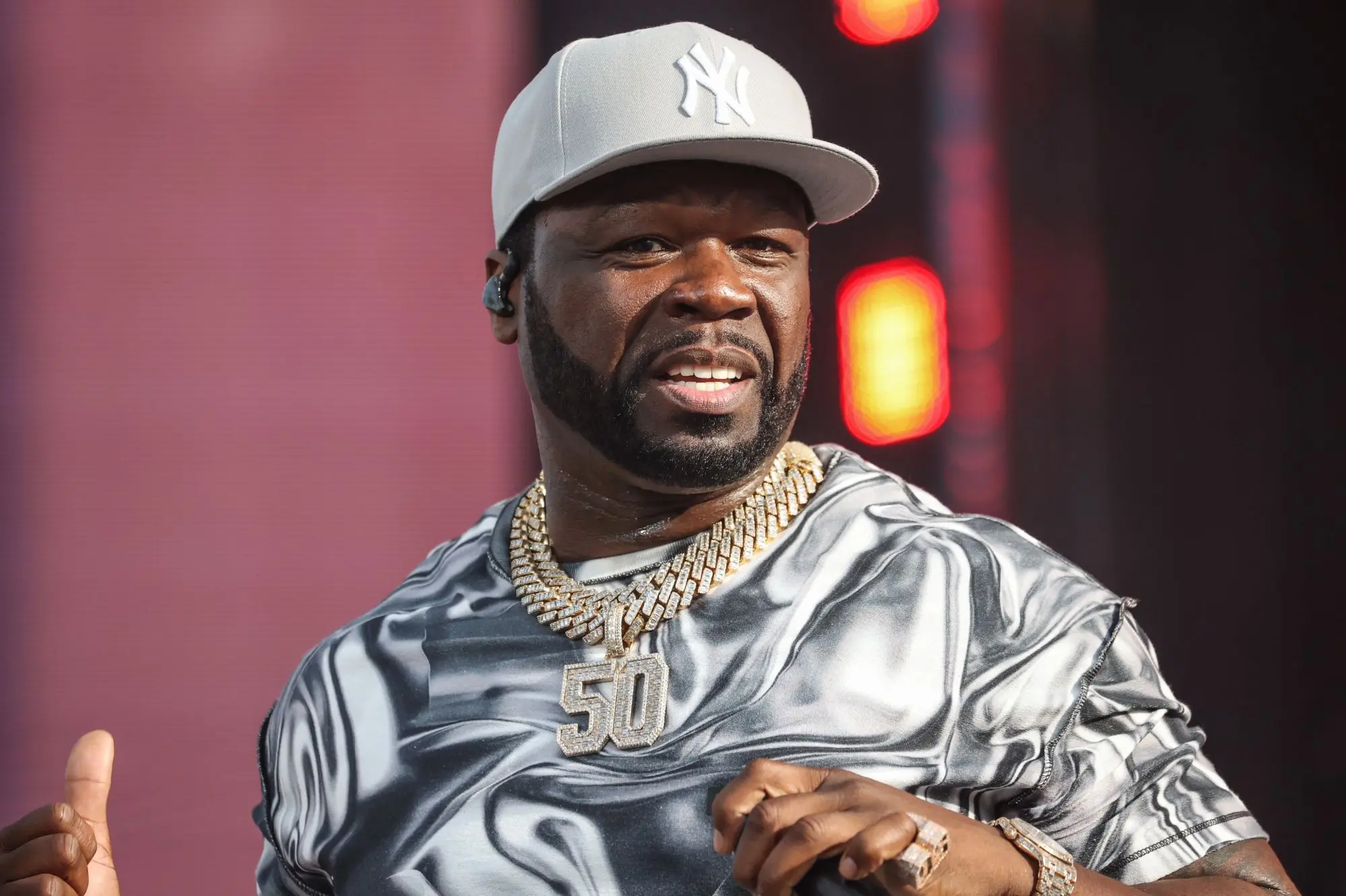 50 Cent performing on stage wearing a silver shirt, New York Yankees cap, and layered diamond chains with a large “50” pendant.