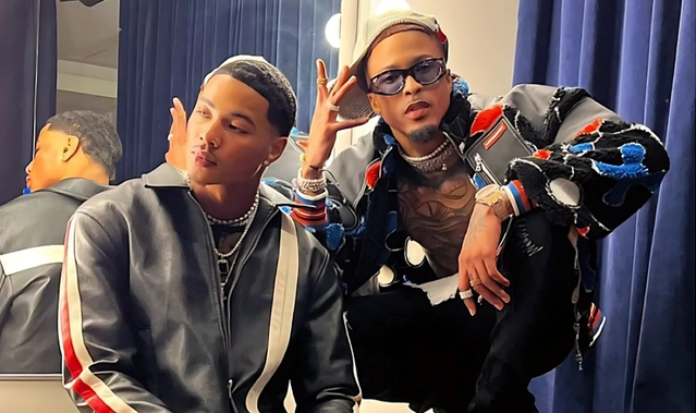 August Alsina wearing sunglasses and a patterned jacket posing next to Zu, who is dressed in a black leather jacket with white and red stripes.