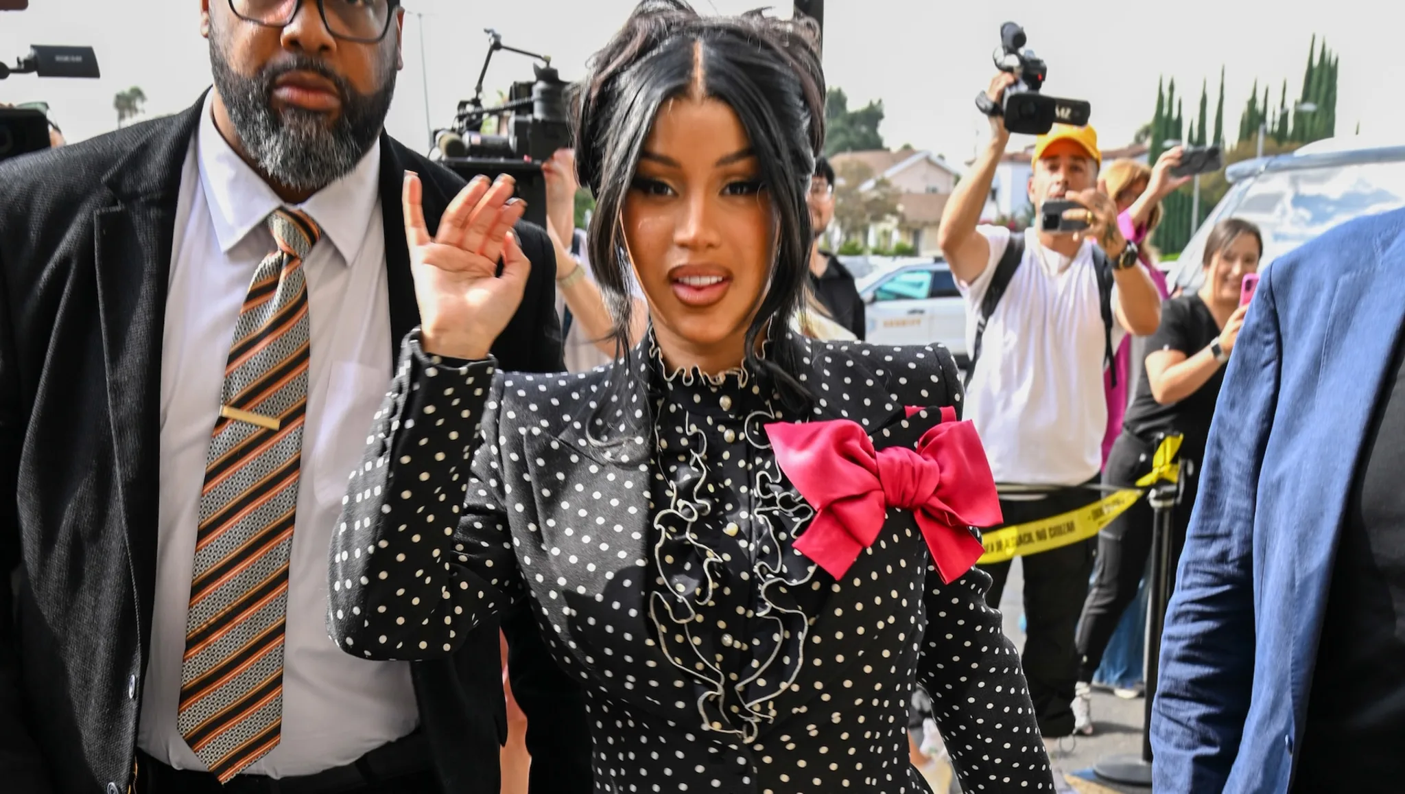 Cardi B wearing a black polka-dot dress with a large red bow, waving to cameras while entering court surrounded by security and media.