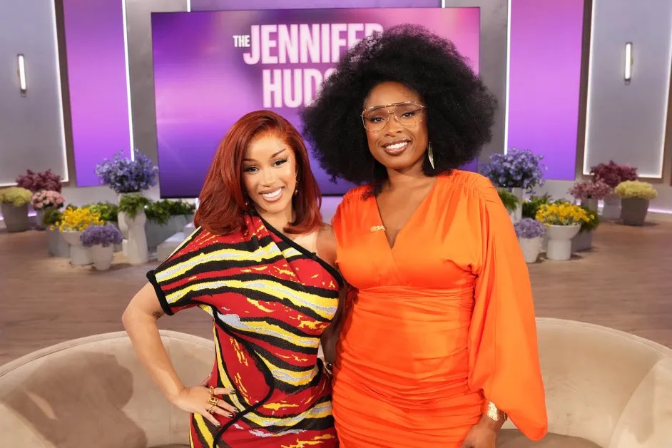 Cardi B in a multicolored dress standing next to Jennifer Hudson in an orange outfit on The Jennifer Hudson Show set.