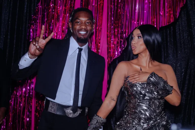Cardi B in a strapless metallic gown and Offset in a black suit posing together at a red carpet event with pink tinsel backdrop.