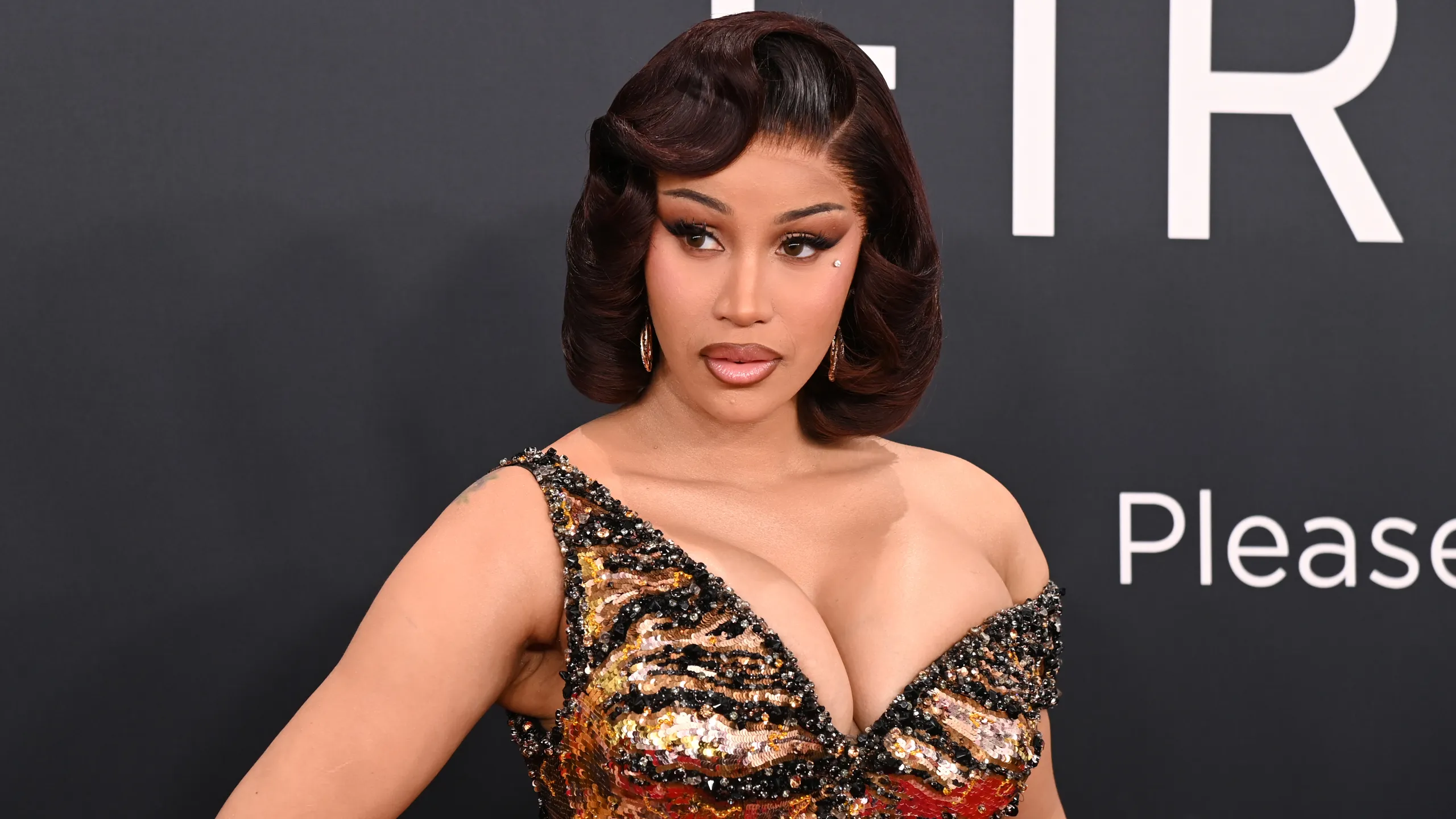 Cardi B wearing a glittering sequin gown with gold, black, and red details, posing on the red carpet with a sleek hairstyle.