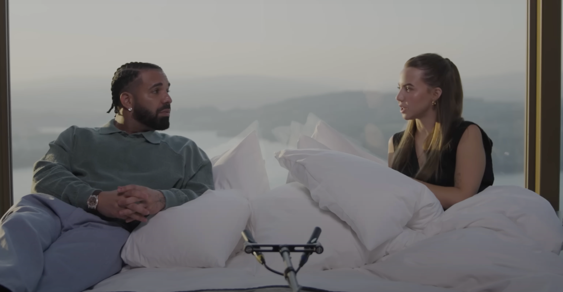 Drake and Bobbi Althoff sitting on a bed during their interview, with pillows in front of a large window overlooking a scenic mountain and lake view.