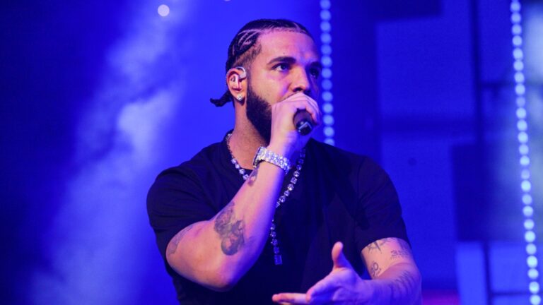 Drake performing on stage in a black shirt, braided hair, diamond chain, and microphone in hand with blue stage lights in the background.