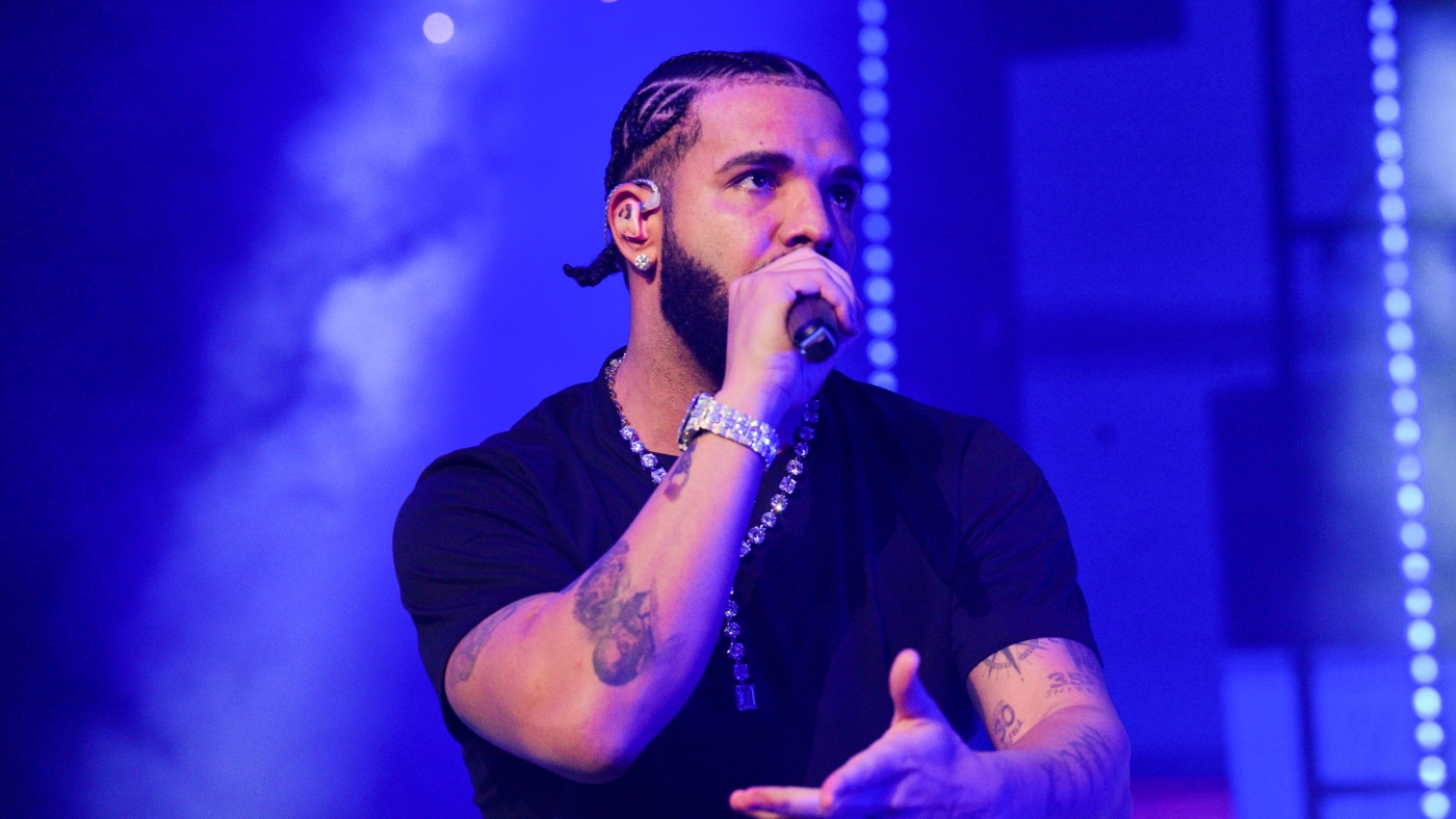Drake performing on stage in a black shirt, braided hair, diamond chain, and microphone in hand with blue stage lights in the background.