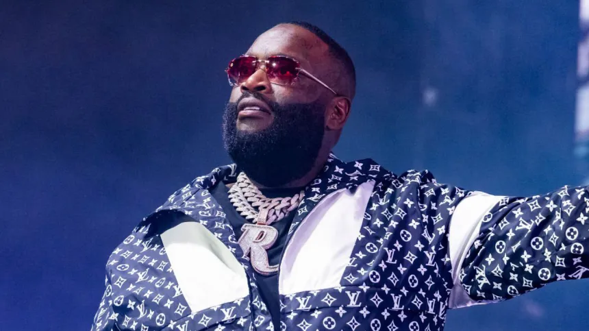 Rick Ross wearing a Louis Vuitton jacket, diamond “R” chain, and red sunglasses while performing on stage.