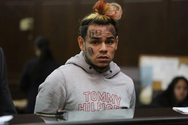 Rapper 6ix9ine standing in court with a Tommy Hilfiger hoodie and tattoos visible on his face.