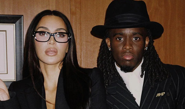 Kim Kardashian wearing glasses and a black outfit posing next to Kai Cenat in a pinstripe suit and black hat.