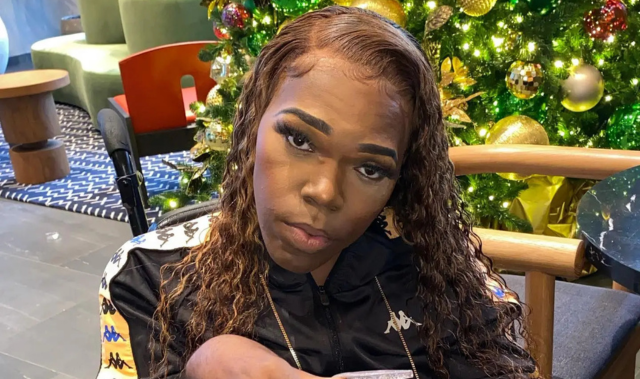 Rolling Ray wearing a black Kappa tracksuit with long wavy hair, seated in front of a Christmas tree with gold and green decorations.