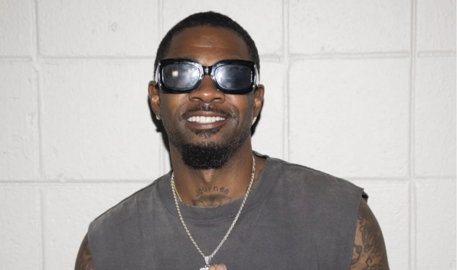 Smiling man in sunglasses and sleeveless shirt posing in front of a white wall