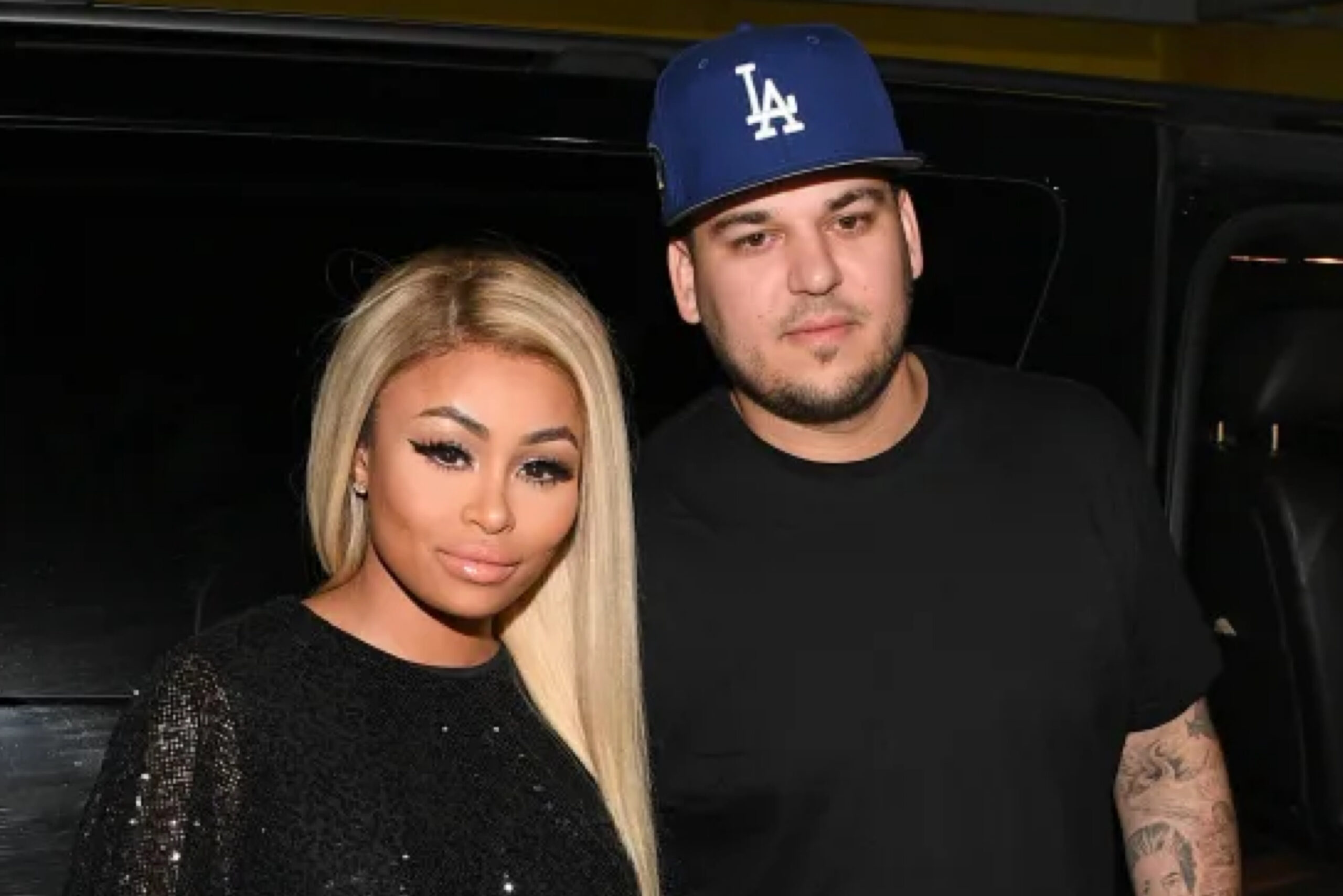 Blac Chyna wearing a black sequined outfit standing beside Rob Kardashian in a black shirt and blue LA hat.