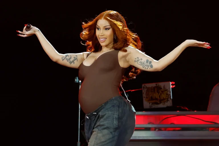 Cardi B performing on stage at the 2025 Global Citizen Festival wearing a brown outfit and smiling with her arms raised.