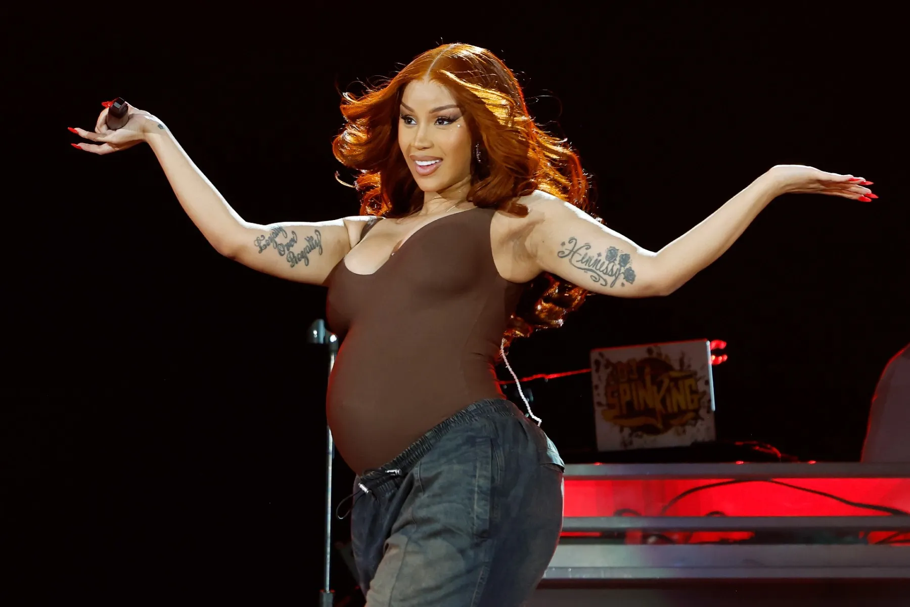 Cardi B performing on stage at the 2025 Global Citizen Festival wearing a brown outfit and smiling with her arms raised.