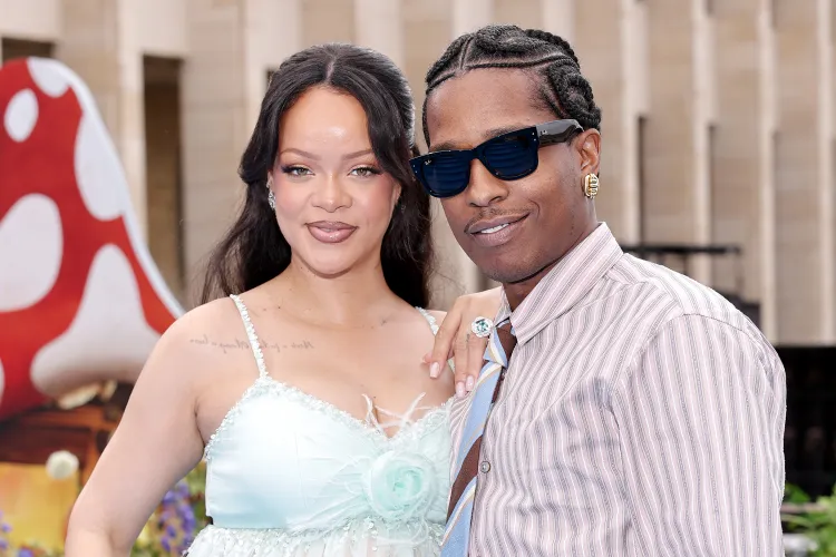 A man and woman stand close together outdoors, smiling for the camera. The woman wears a light blue dress, and the man wears sunglasses and a striped shirt with a tie.