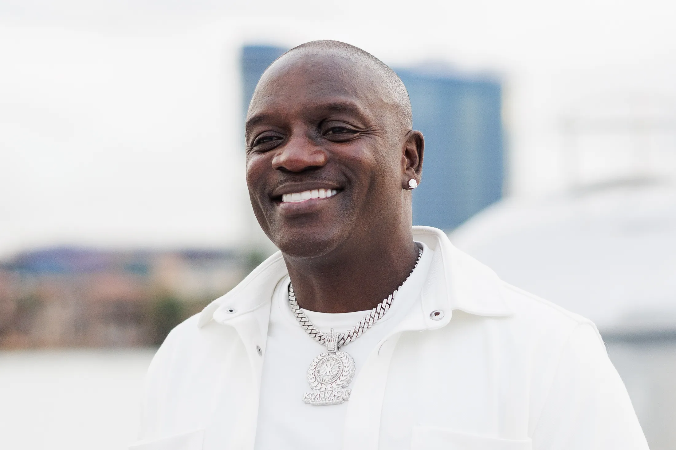 Akon wearing a white shirt and jacket with a diamond chain, smiling outdoors with a blurred cityscape background.