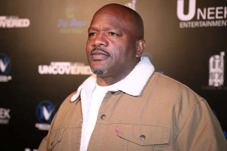 A man in a brown sherpa-collar jacket stands in front of a black step-and-repeat backdrop at an entertainment industry event.
