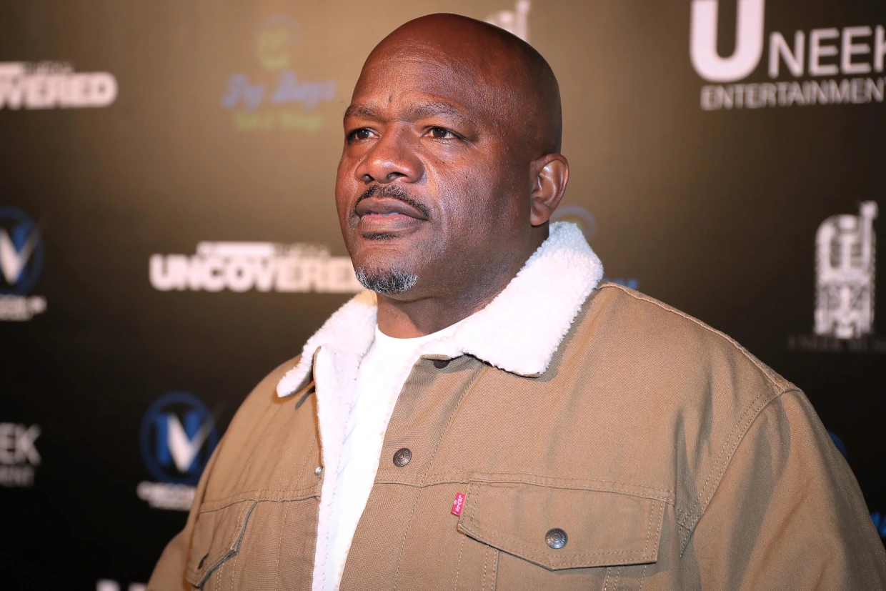 A man in a brown sherpa-collar jacket stands in front of a black step-and-repeat backdrop at an entertainment industry event.