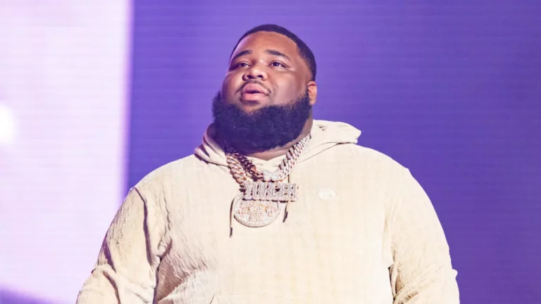 Man with beard wearing a cream hoodie and gold chains standing on stage with purple lighting in the background.