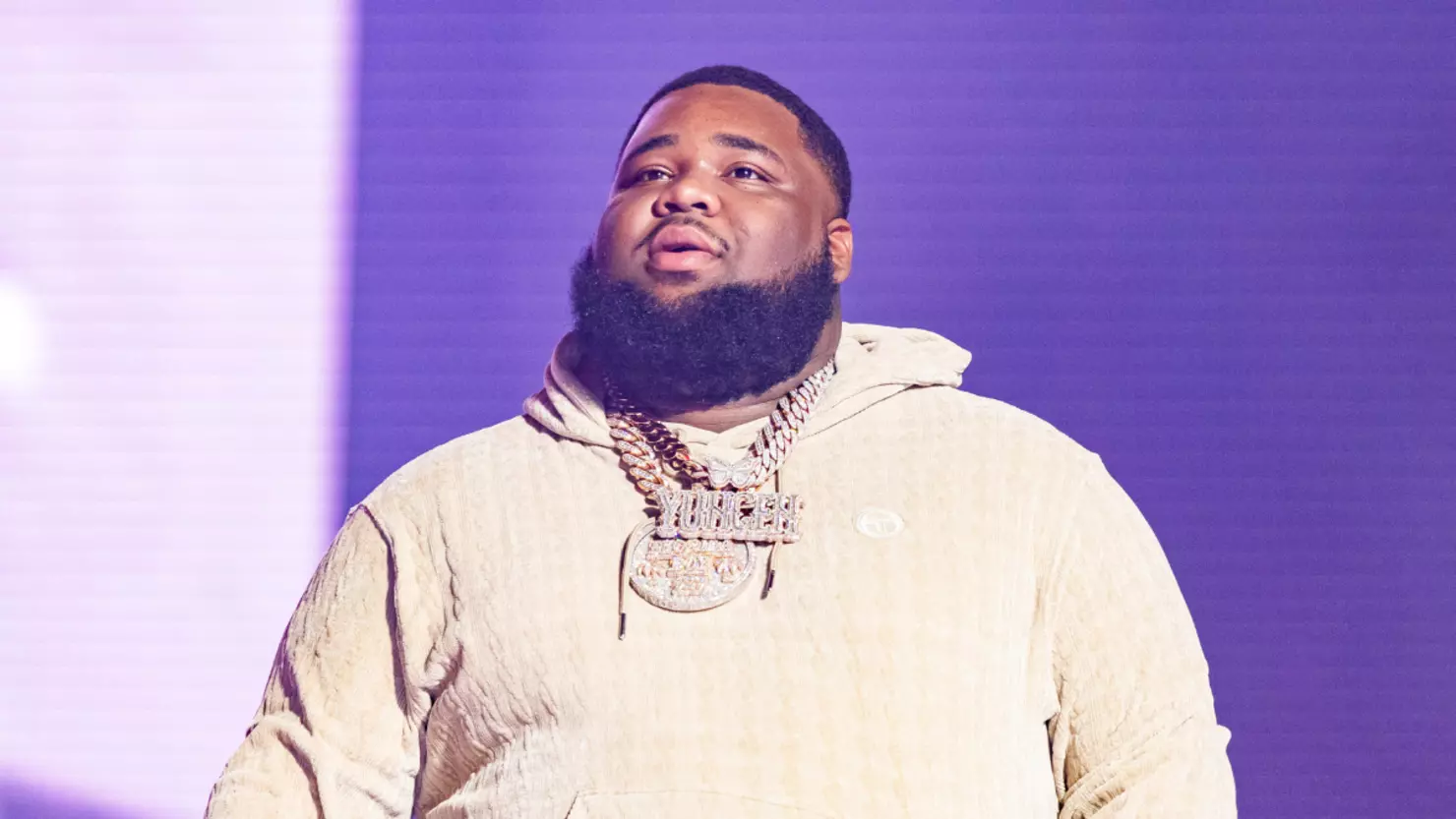 Man with beard wearing a cream hoodie and gold chains standing on stage with purple lighting in the background.