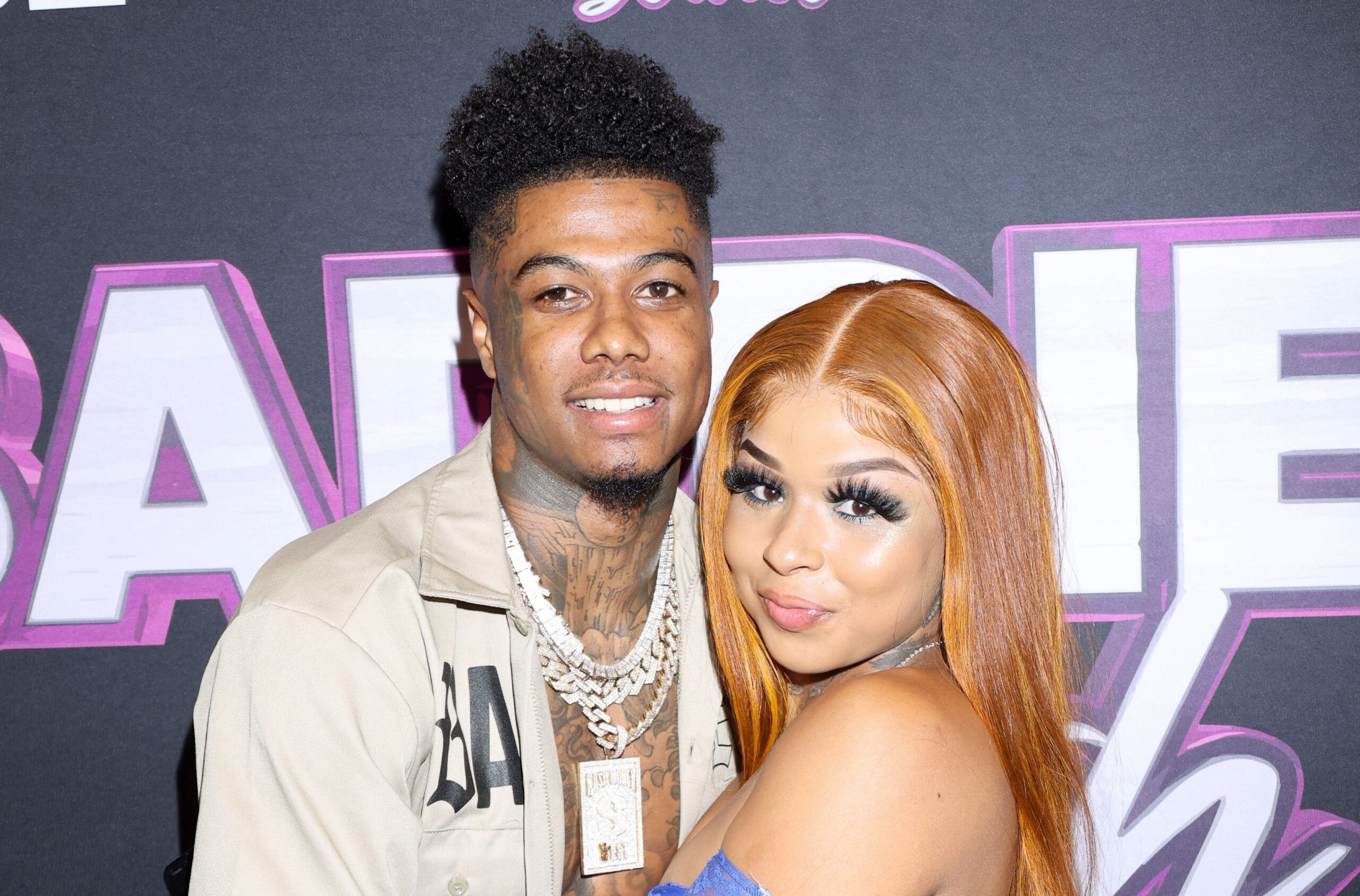 Blueface wearing layered diamond chains and a beige jacket standing beside Chrisean Rock with orange hair and a blue outfit at a red carpet event.