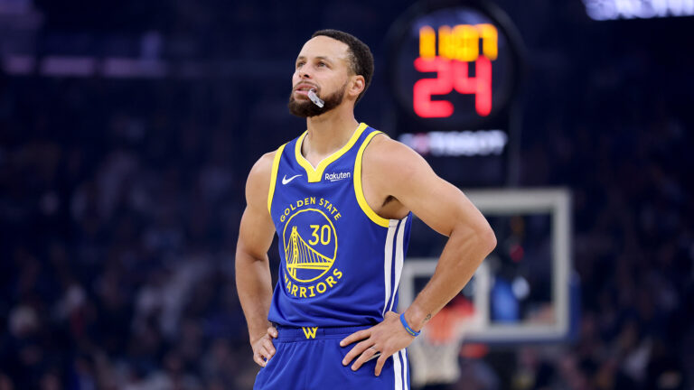 Basketball player wearing a blue and yellow jersey standing with hands on hips during a game, with a lit shot clock behind him.