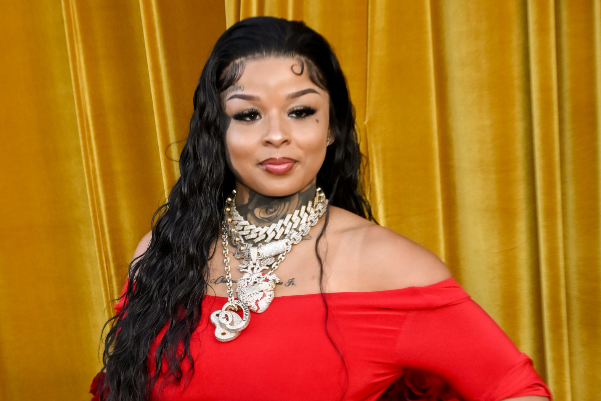 Woman with long wavy hair and layered jewelry wearing a red off-the-shoulder dress in front of a gold