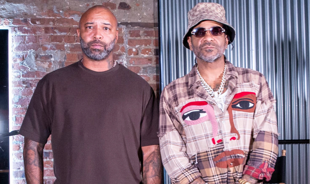 Two men standing together indoors, one wearing a plain dark T-shirt and the other wearing patterned streetwear with sunglasses and a bucket hat.