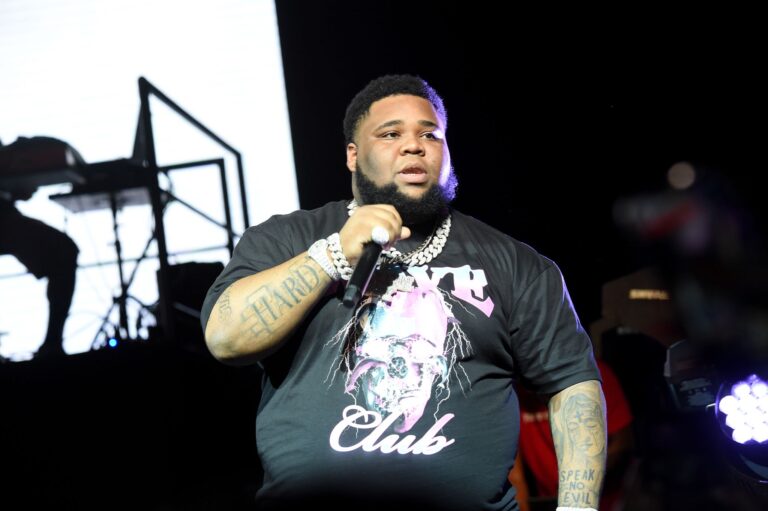 Rod Wave performing live on stage wearing a beige hoodie and diamond jewelry under purple lighting.