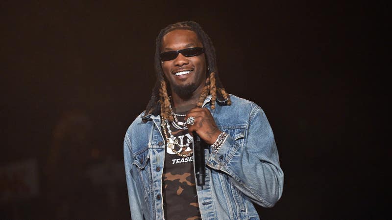 Man in a denim jacket and sunglasses smiling on stage while holding a microphone.