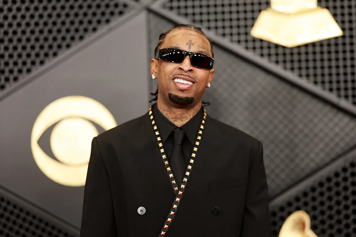 Man wearing black suit, sunglasses, and braided hairstyle posing on the red carpet in front of music awards backdrop.