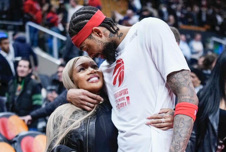 Basketball player in a white team shirt holding and smiling down at a woman with long blonde hair, sharing an affectionate moment courtside with fans in the background.