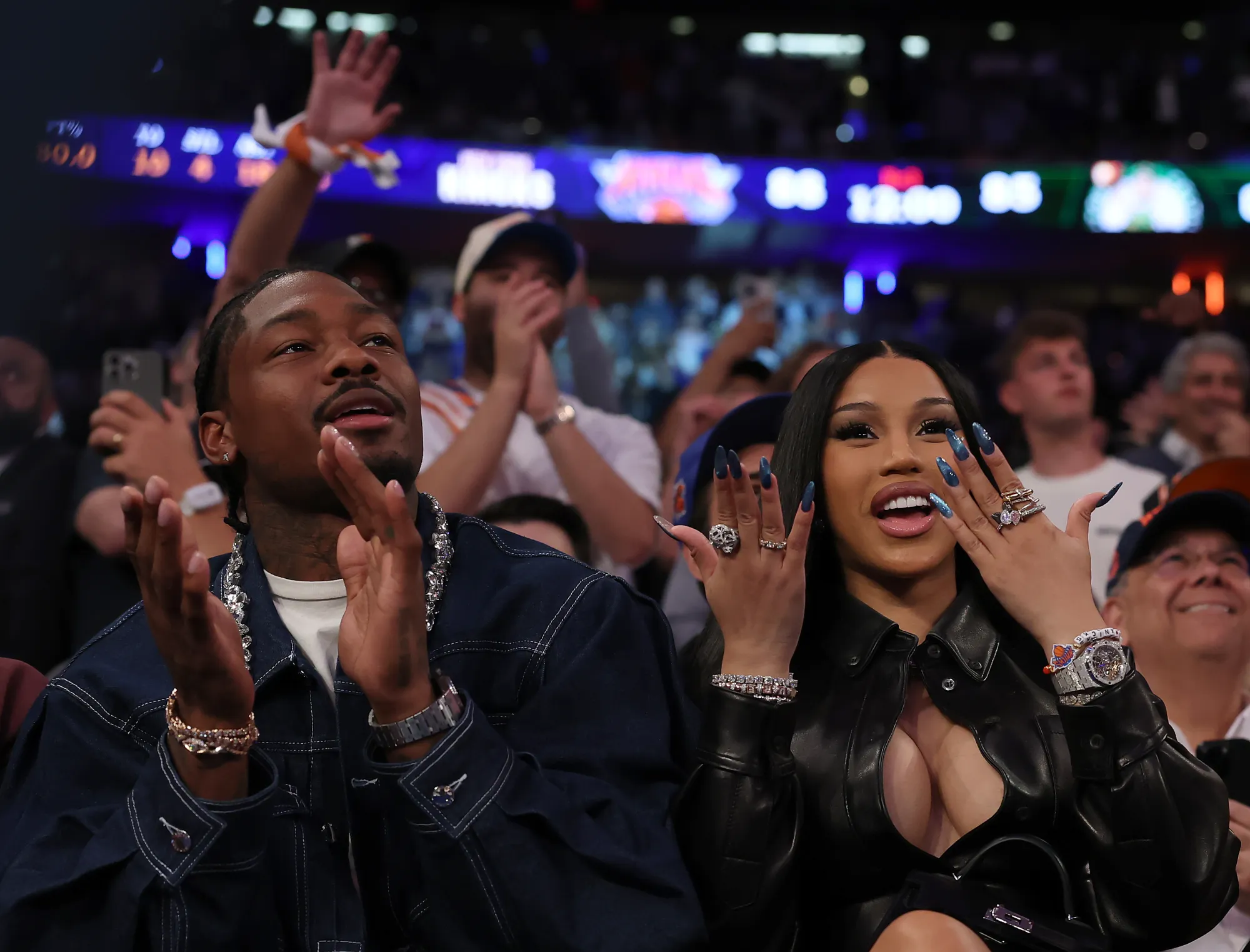 Man and woman sitting courtside at a basketball game, cheering and smiling with excited reactions while wearing stylish outfits and jewelry.