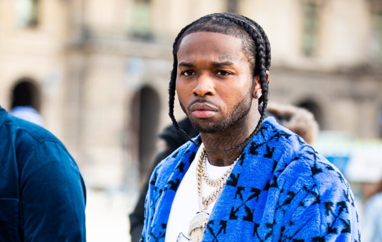 Man with braided hair wearing a bright blue patterned coat and gold jewelry while standing outside in an urban area.